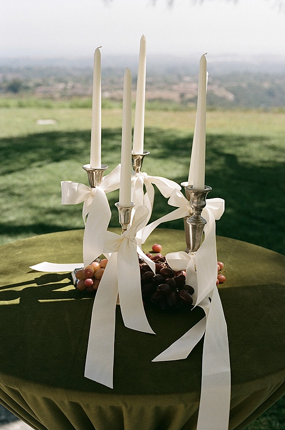 Wedding table centerpiece with taper candles in silver candlesticks, ivory ribbons, and grapes on a green tablecloth set on a lawn with hills