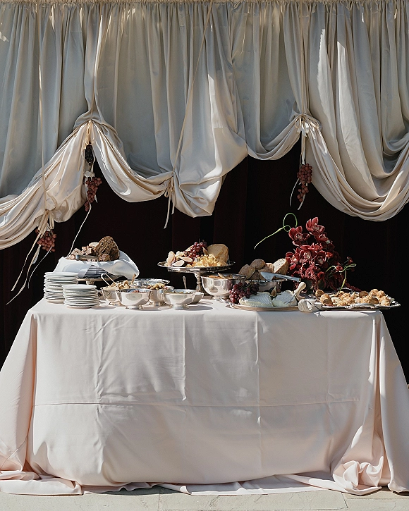 Wedding dessert table with a wedding grazing table spread of bread, cheese and grapes on white linens, set against a black backdrop and stone floor