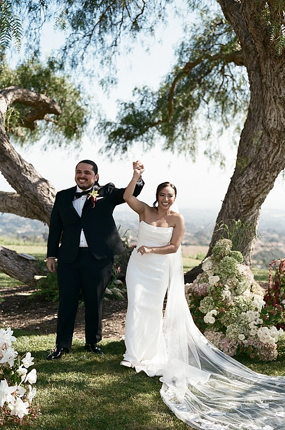 Recessional moment as newlyweds walk hand in hand cheering, bride in strapless dress and long veil beside groom in tuxedo on a lawn aisle with flowers under large trees and blue sky
