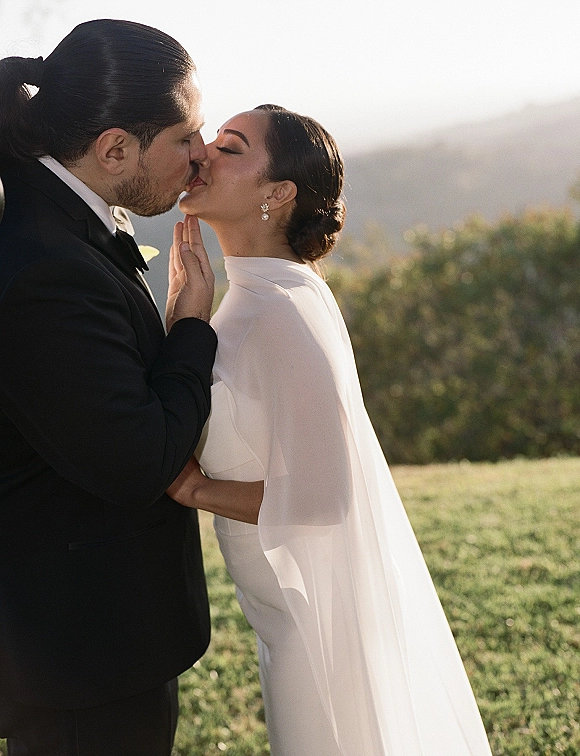 Wedding kiss portrait of bride and groom kissing, her veil and sheer cape flowing as he cups her face on a hillside with mountains beyond