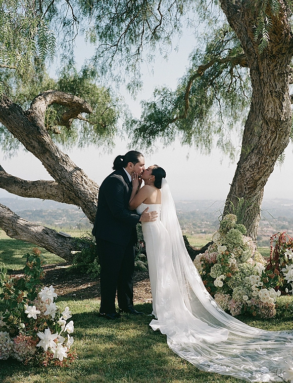 Wedding kiss portrait of bride and groom kissing, her long veil flowing over grass beneath a large tree with floral greenery accents