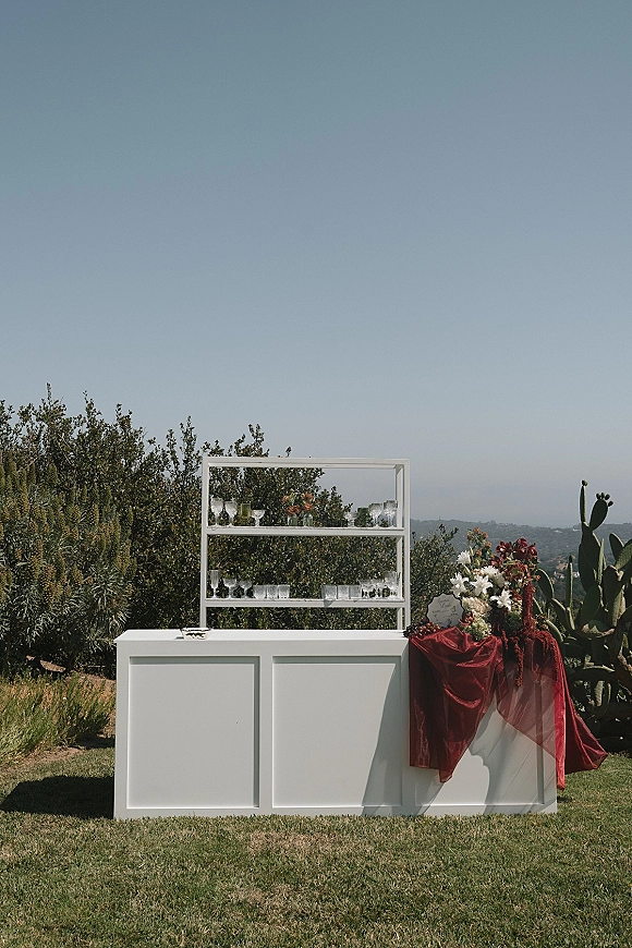 Wedding bar setup with a white outdoor wedding bar, back bar shelving, glassware, and floral arrangement on a lawn with cactus backdrop