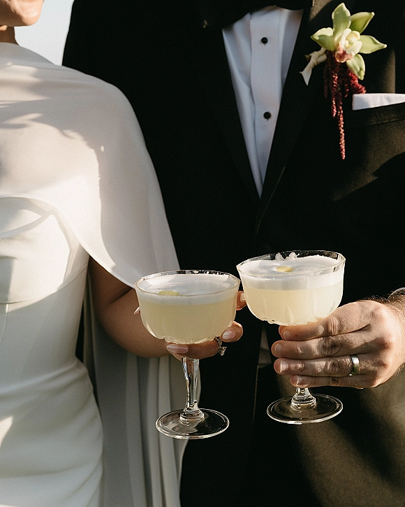Wedding toast as newlyweds clinking glasses of pale yellow cocktails with lemon garnish, bride in strapless dress and groom in tuxedo in soft light