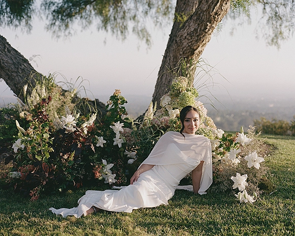 Bridal portrait of a bride reclining on grass in a white gown with cape sleeves, beside a lush lily and hydrangea floral installation under a large tree