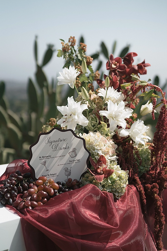 Signature drink sign with wedding signature drinks menu beside white lilies, orchids and grapes on burgundy drape, with cacti and sky behind