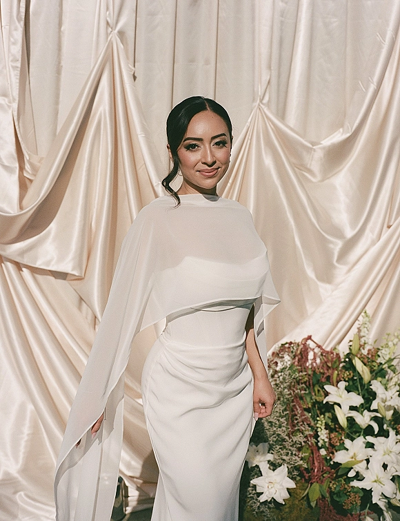 Bridal portrait of a bride in a minimalist wedding dress with cape sleeves, standing by satin drape backdrop with lilies and greenery