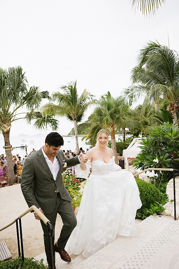Wedding recessional as bride and groom walking hand in hand down stone steps, ocean and palm trees behind, guests watching nearby