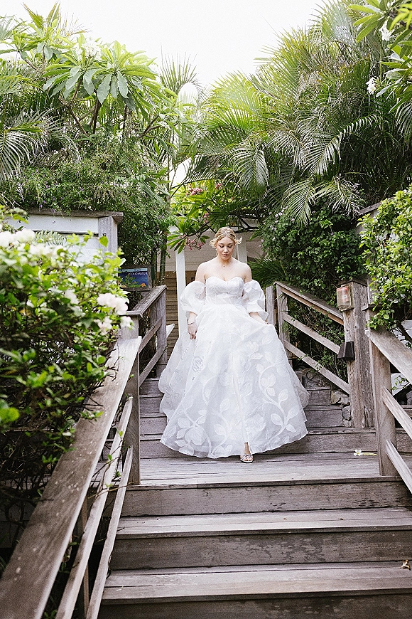 Bridal portrait of a plus size bride walking on a wooden boardwalk in a strapless embroidered ball gown with off-shoulder sleeves amid palms