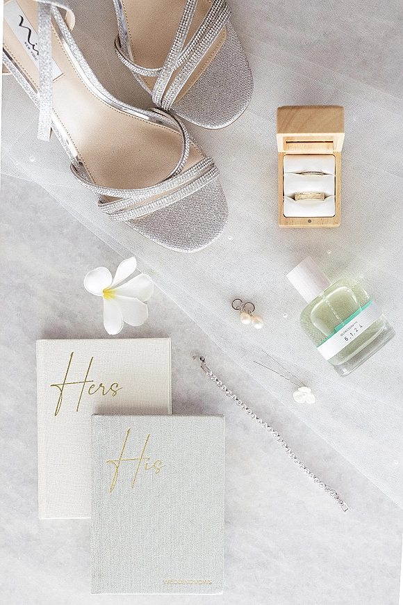 Wedding details flatlay with bridal accessories flatlay, silver strappy heels, velvet ring box, vow books, jewelry, perfume and white flower on stone surface
