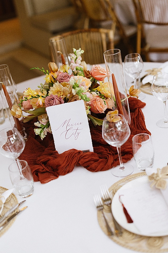 Reception tablescape with wedding table centerpiece of peach and mauve roses, bud vases and taper candles on a rust gauze runner over white linen tablecloth