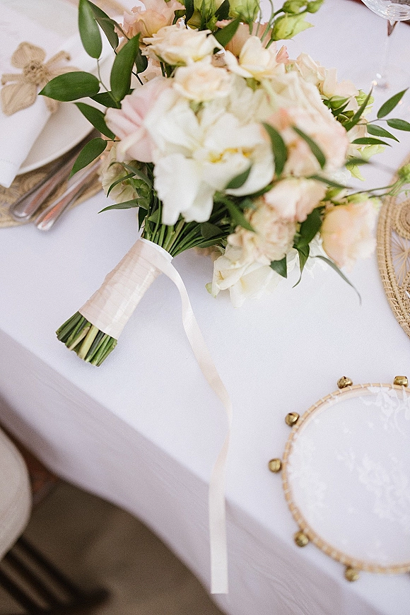 Bridal bouquet of blush and white bouquet blooms with greenery on a white-tablecloth place setting beside plates, glassware and a tambourine