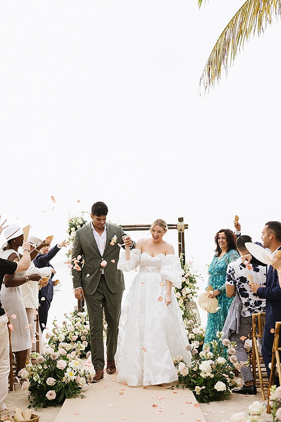 Wedding recessional as newlyweds walk the aisle while guests toss rose petals, under a simple arch with palm fronds and bright sky overhead