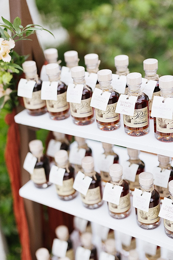 Wedding favors of mini liquor bottle favors with personalized favor tags tied in twine on a wooden stand, framed by garden greenery
