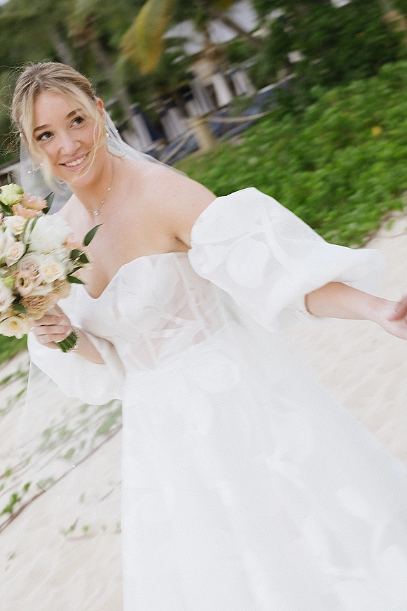 Bridal portrait of a bride holding bouquet in an off the shoulder wedding dress, smiling on a boardwalk with palm trees and sand behind