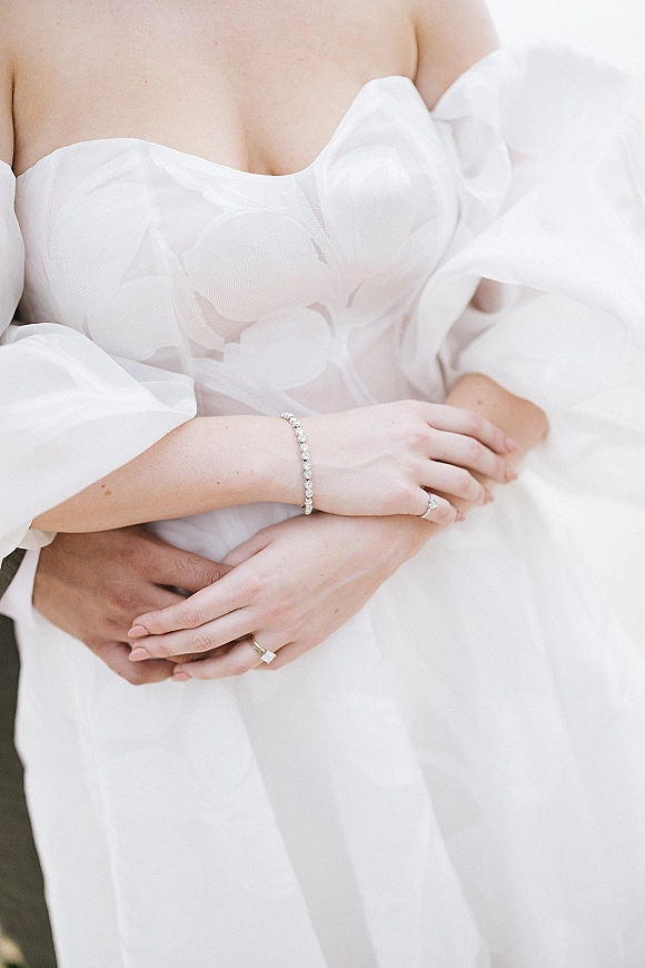 Bridal gown close-up showing an off the shoulder wedding dress with floral appliqué bodice, sheer puff sleeves, and diamond bracelet rings on hands
