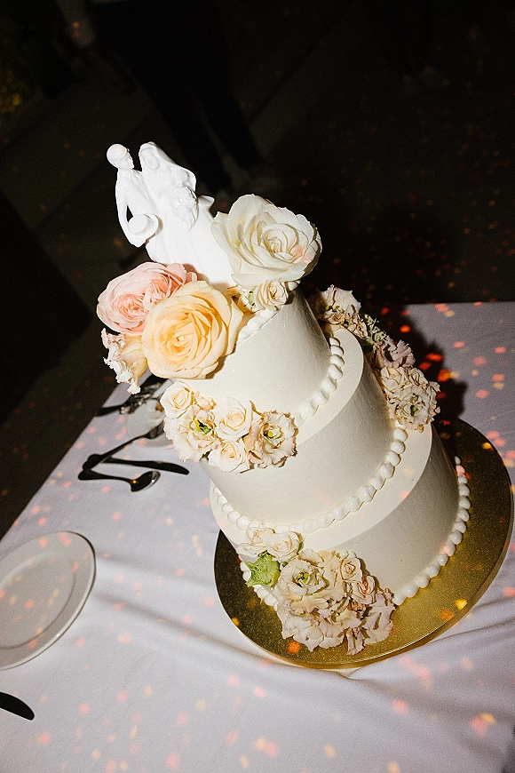 Wedding cake with white icing and beaded border, topped with fresh roses and figurine on a gold board against a dark bokeh backdrop