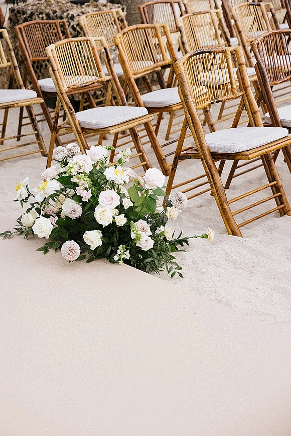 Ceremony seating with wedding chair decor, wood chairs with white cushions lined on sand beside low rose and greenery aisle florals