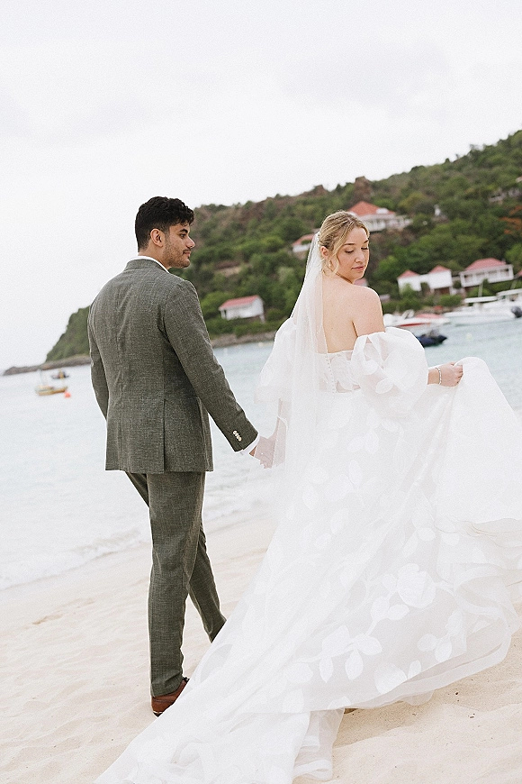 Couple portrait of beach wedding couple holding hands on a sandy shoreline, bride in veil and dress train, boats and villas behind