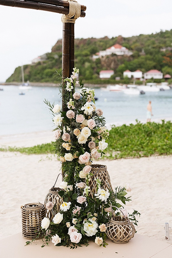Wedding ceremony arch with bamboo and rope ties, blush roses and greenery, plus a rattan lantern accent on sandy beach by the ocean
