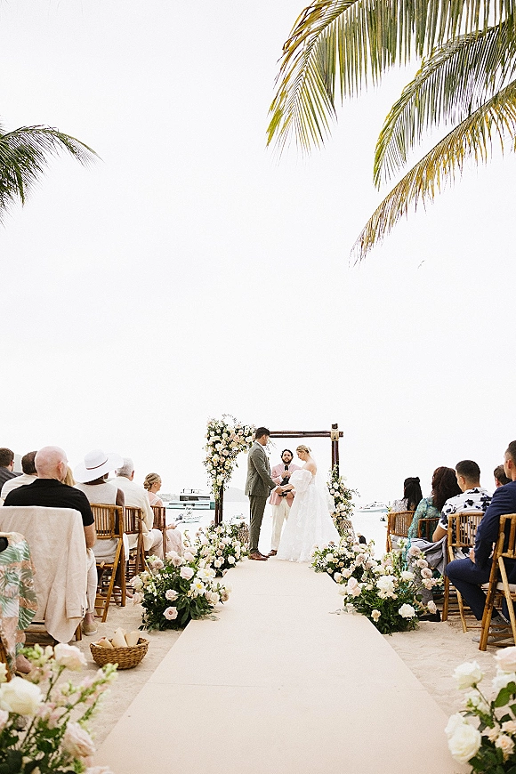 Wedding ceremony on a sandy beach with bride and groom at a floral arch, white roses and greenery lining the aisle, ocean and boats behind
