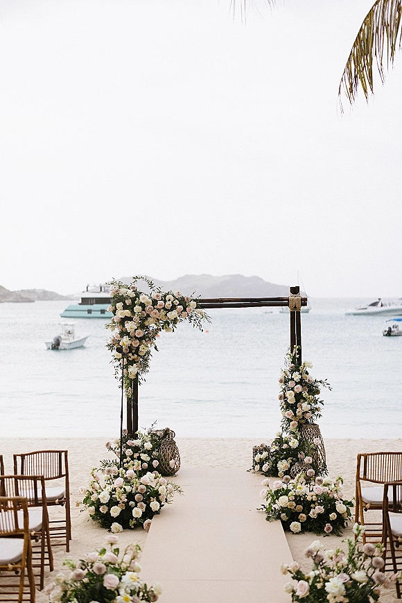 Beach ceremony setup with a wedding floral arch of blush roses, aisle runner, wooden chairs, and woven lanterns facing the ocean