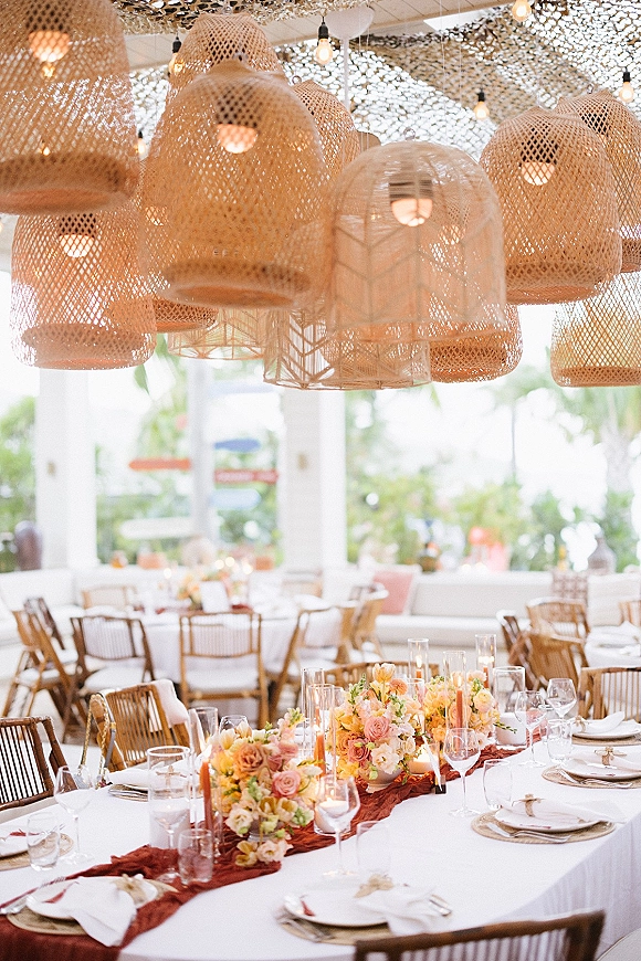 Reception tablescape with wedding table centerpiece, taper candles and lush florals under rattan pendant lights in an open-air pavilion with columns