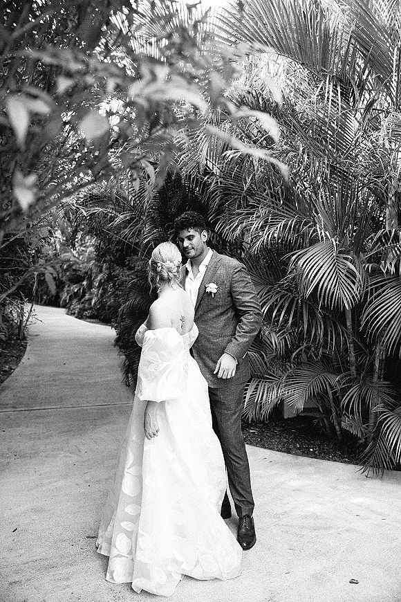 Couple portrait in black and white wedding portrait style, bride and groom embrace with a forehead kiss on a palm-lined garden path