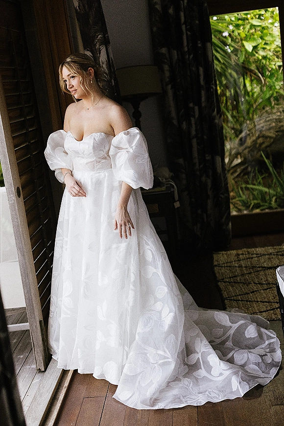 Bridal portrait of a bride in an off the shoulder wedding dress, standing by window light with wooden shutters and greenery outside