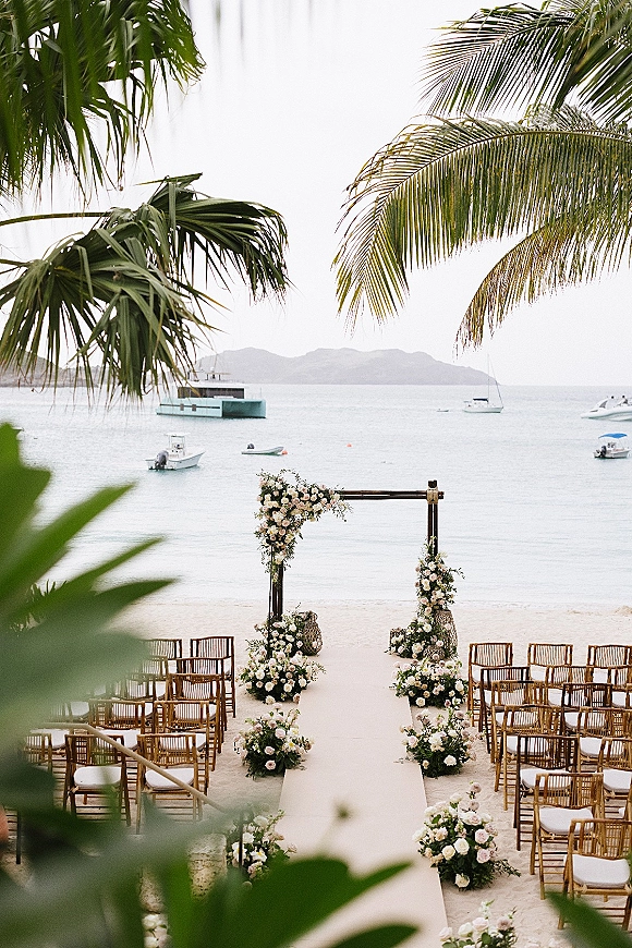 Beach ceremony setup with a floral wedding arch, white aisle runner, lanterns and chairs on sand facing the ocean and hills