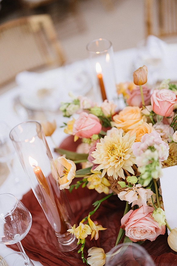 Reception tablescape with wedding table centerpiece of pastel roses, dahlias and tulips, taper candles in glass, and wine glasses on a white cloth