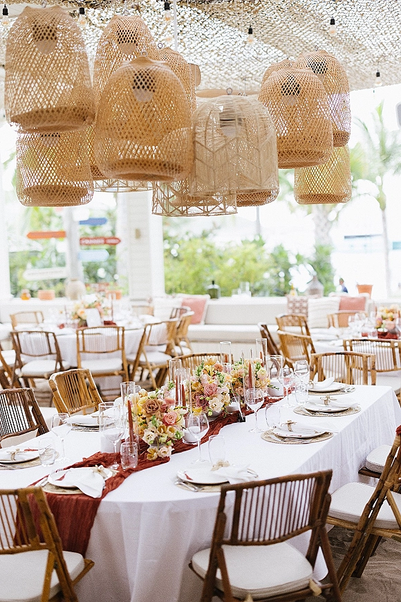 Reception tablescape with wedding head table decor featuring rust runner, low florals, tapered candles, and rattan chairs under string lights in pavilion
