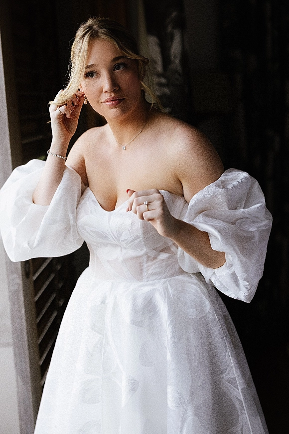 Bridal portrait of a bride by window light adjusting her pearl drop earrings in an off-the-shoulder puff sleeve gown indoors