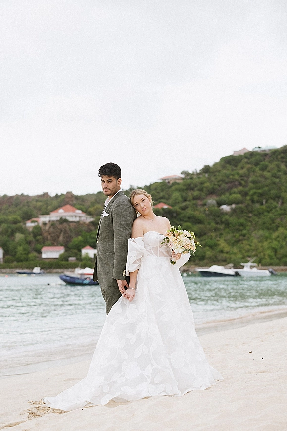 Couple portrait of bride in strapless dress holding a bouquet, leaning on groom in suit, with ocean, boats and hills behind