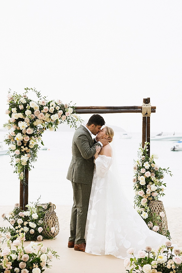 Wedding kiss portrait of bride in an off-shoulder dress and groom under a rose and greenery arch on a beach with ocean and boats behind