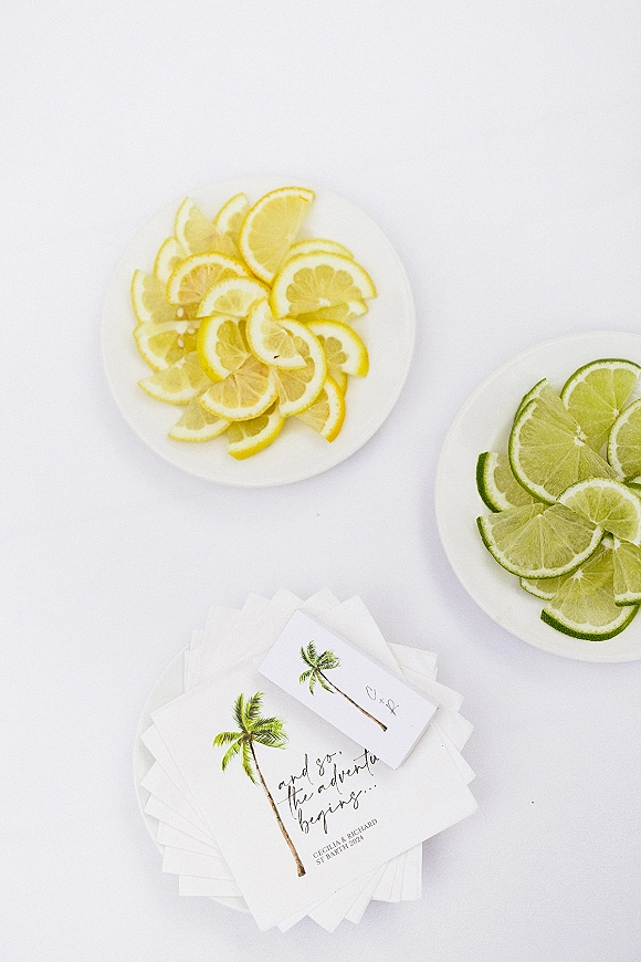 Wedding invitations with a palm tree illustration and script lettering, styled with lemon and lime slices on white plates over a white surface