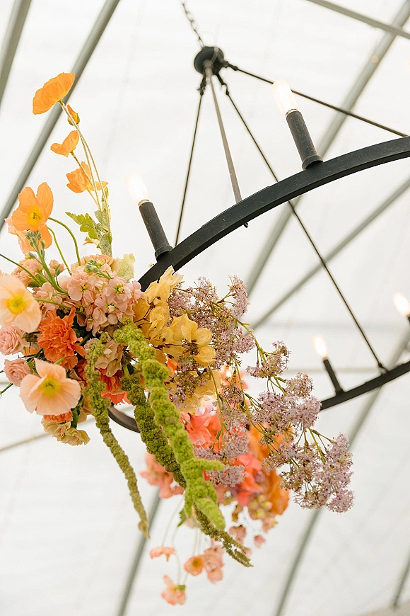 Floral chandelier with hanging wedding flowers in orange, peach, yellow, and lavender blooms on a black candle-light fixture under a glass ceiling