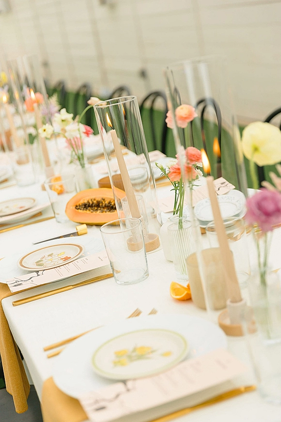 Reception tablescape with pastel bud vases and taper candles on a long banquet table, gold flatware, patterned plates, and green velvet chairs