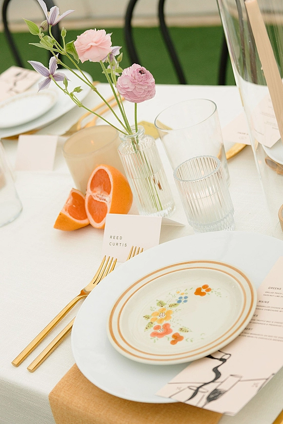 Reception tablescape wedding place setting with striped bud vase of ranunculus and clematis, orange slices, gold flatware on white tablecloth outdoors
