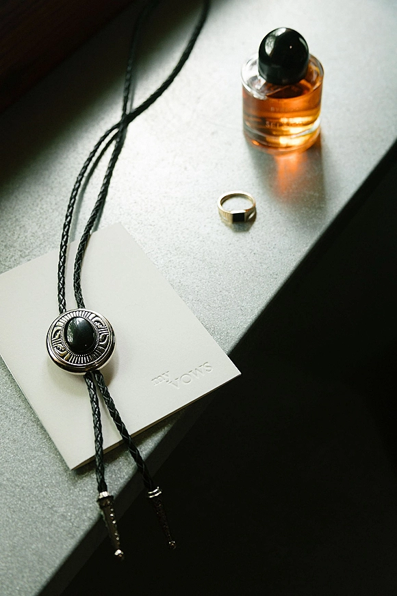 Groom details wedding flat lay with gold wedding band, cologne bottle, bolo tie and gift box on a stone countertop in window light