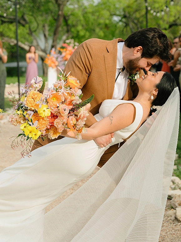 Wedding kiss portrait of groom dipping bride as she holds a colorful bouquet, veil flowing in a garden with string lights and guests