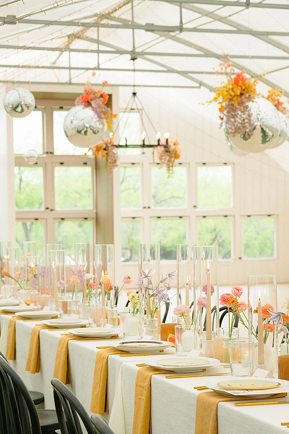 Reception tablescape with long head table decor featuring a mustard runner, bud vase flowers, taper candles, and string lights under a glass ceiling