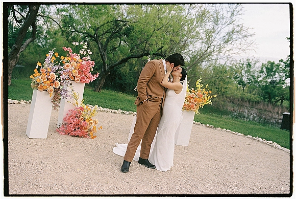 Wedding kiss portrait of bride and groom kissing, her veil flowing as she holds his face on a gravel path framed by floral pedestals