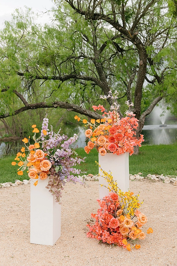 Ceremony floral pillars with colorful roses and wildflowers on white pedestal stands, set by a pond and fountain amid trees