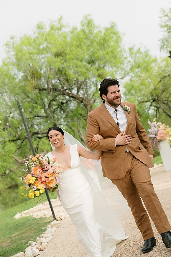 Couple portrait of bride and groom walking on a gravel path, bride in a white dress with veil holding a colorful bouquet among trees