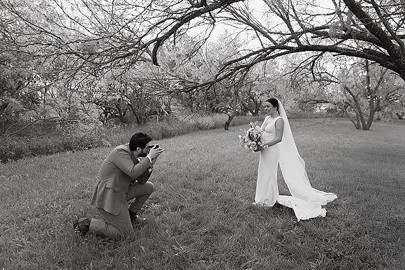 Bridal portrait of a bride holding bouquet, smiling in a simple sheath wedding dress with long veil, as a photographer kneels on a wooded lawn