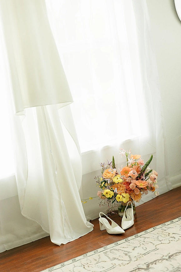 Bridal details with wedding dress and shoes, colorful bouquet in a glass vase and white heels by a sunlit window with sheer curtains