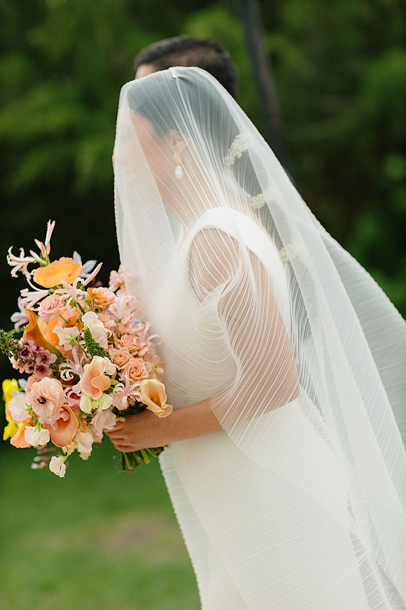 Bridal portrait of a bride with veil draped over her face, holding an orange and blush bouquet in a strapless gown outdoors amid blurred trees