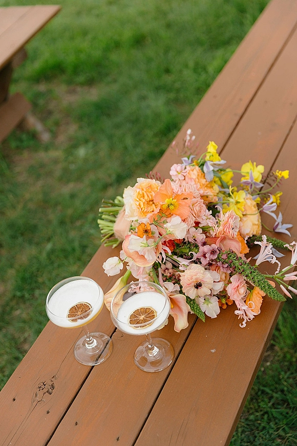 Bridal bouquet with garden style blooms in pastel peach, orange, and yellow beside coupe champagne cocktails with dried citrus on a wooden bench