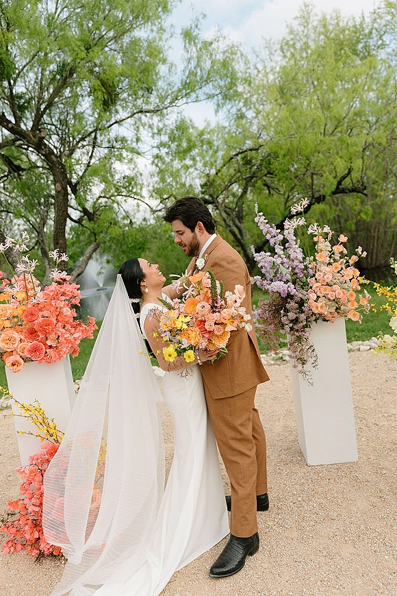 Couple portrait of bride looking up at groom in tan suit, holding a colorful bouquet with veil flowing by pond and floral plinths