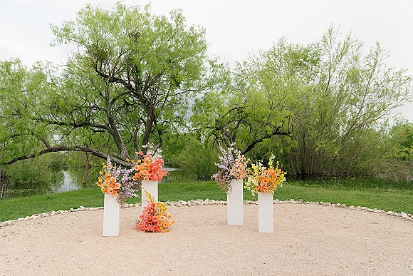 Ceremony floral pillars with wedding aisle pedestal flowers on white plinths, lush mixed blooms on gravel by a pond under trees